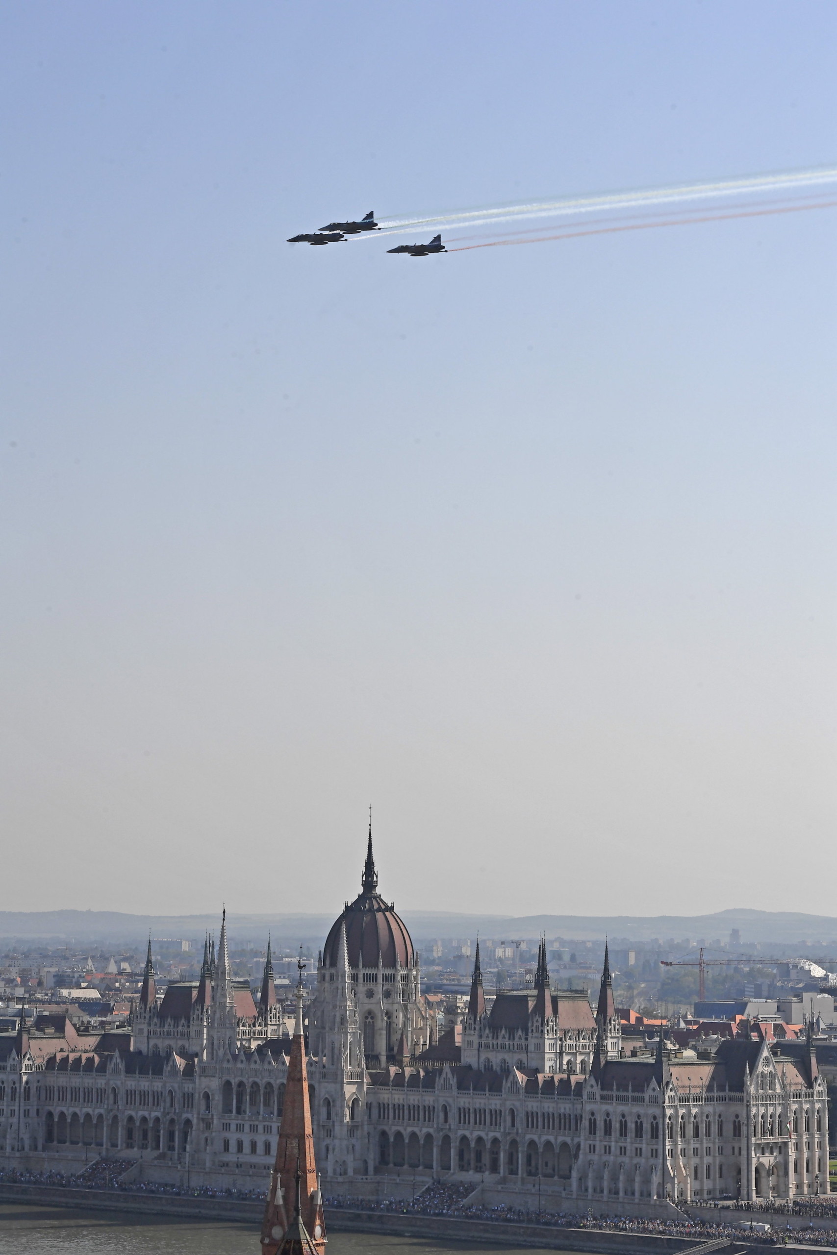 A Magyar Honvédség JAS-39 harcászati repülőgépeinek díszelgő áthúzása nemzeti színű füsttel a Duna felett, a háttérben az Országház épülete.