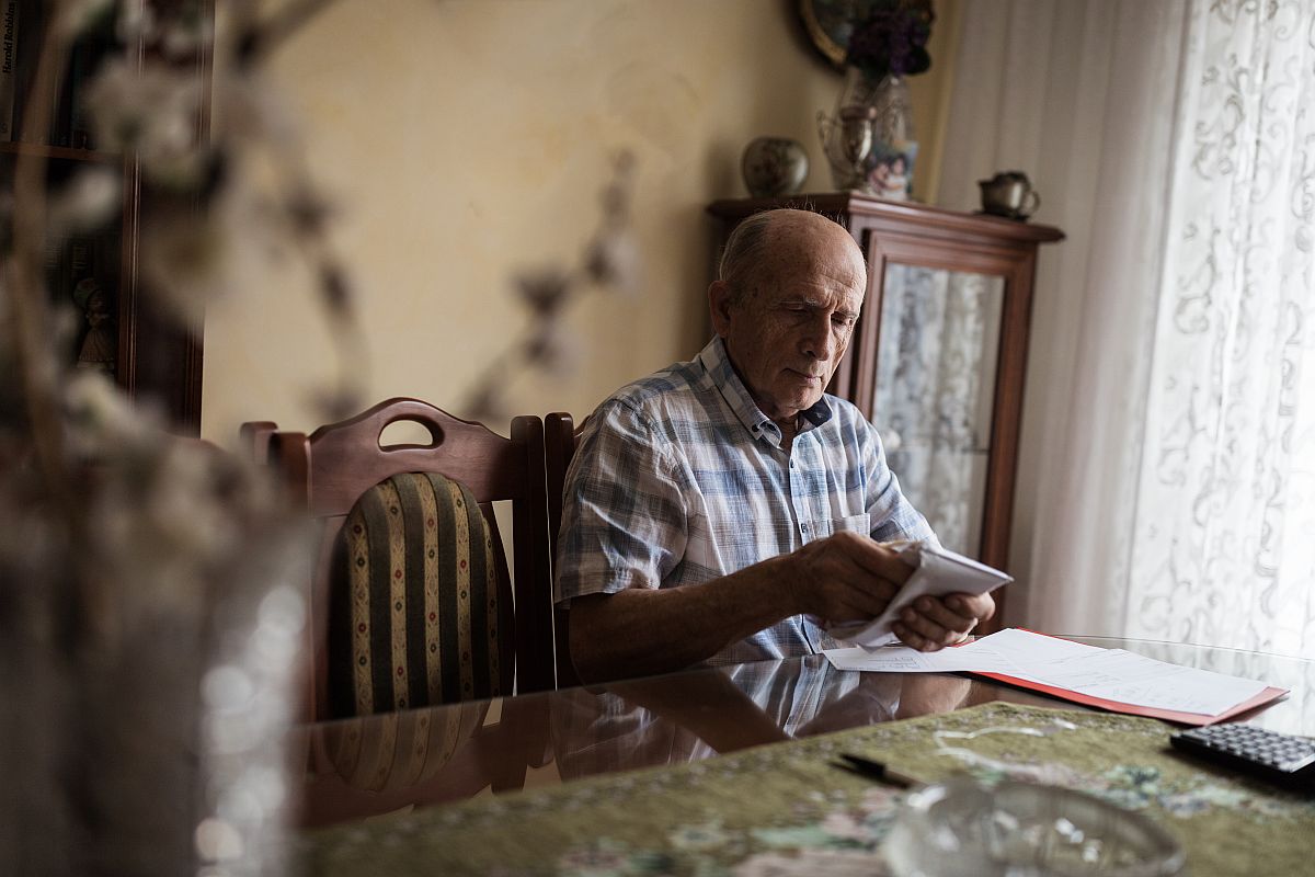 Egy nyugdíjas egy borítékot tart a kezében egy asztalnál ülve a szobában.