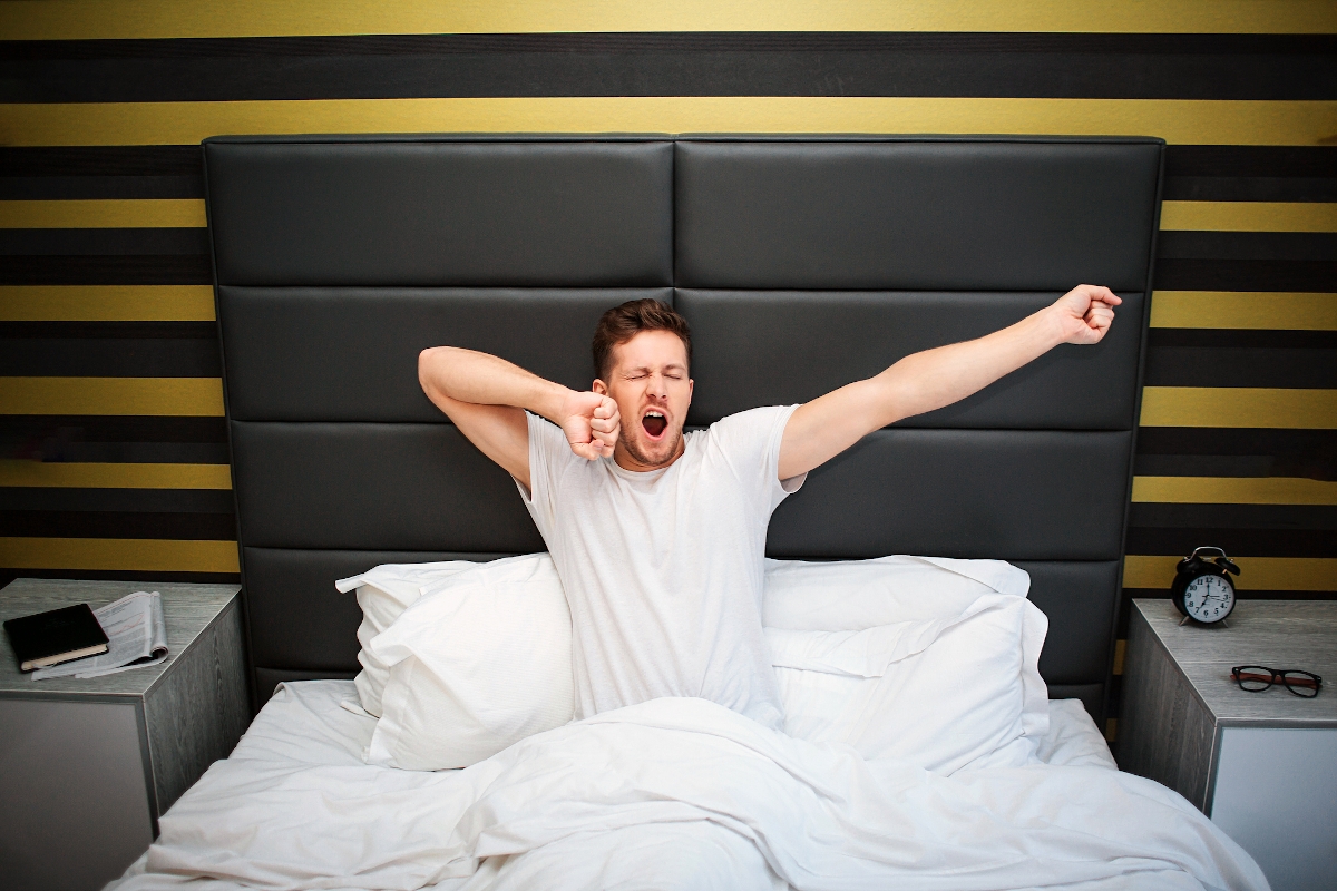 Fiatal férfi ül az ágyban, és nyújtózkodva ásít