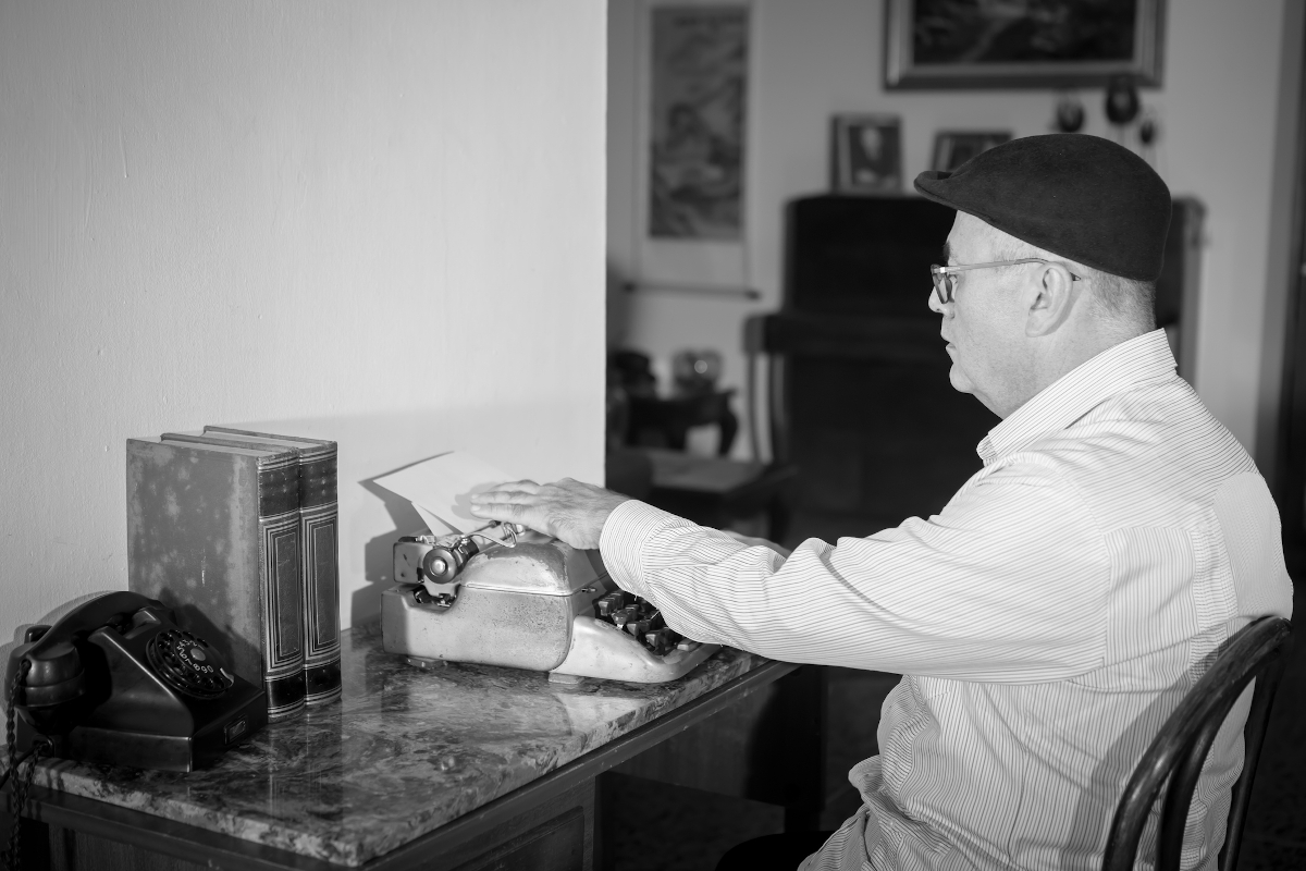 irogeppel_ir_ - Startlap Régies hangulatú, fekete-fehér felvétel egy újságíróról vagy íróról, aki régi mechanikus írógép mellett ül. Az asztalon antik telefon látható, a szereplő korabeli öltözéket és svájcisapkát visel.