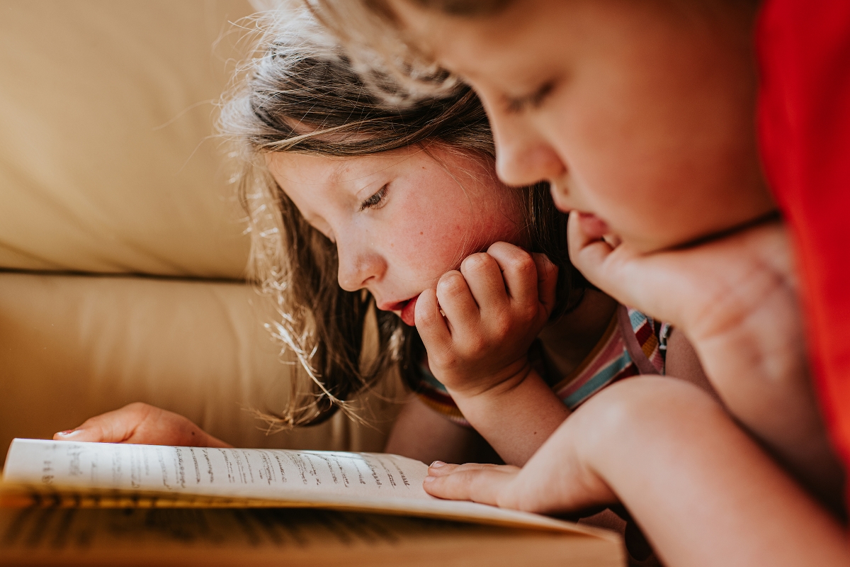 olvas_children lie on their tummies and read the same book together - Startlap Két gyerek osztozik egy könyvön. Hasra fekszenek, a fejüket a kezükre hajtják, és együtt olvassák a könyvet.