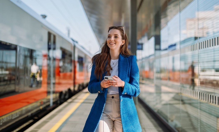 Egy mosolygó utas mobiltelefonnal a kezében vonatok között a peronon.