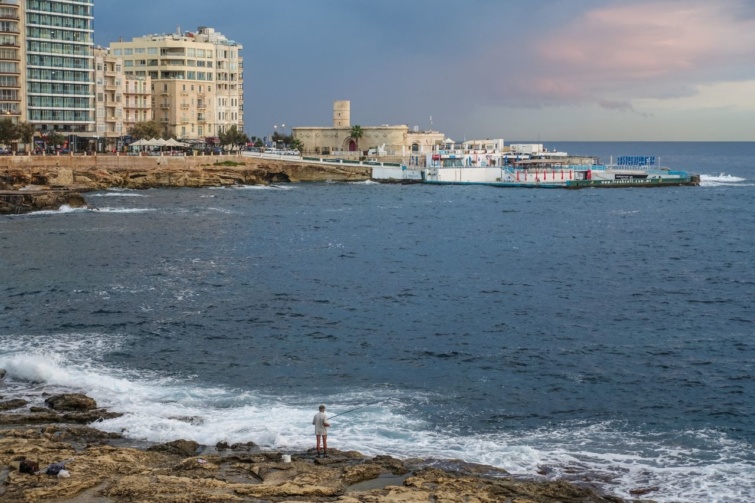 Napkeltekor a sziklás partról egy tengerben halászó férfi látható a máltai San Ġiljan melletti Sliemában.