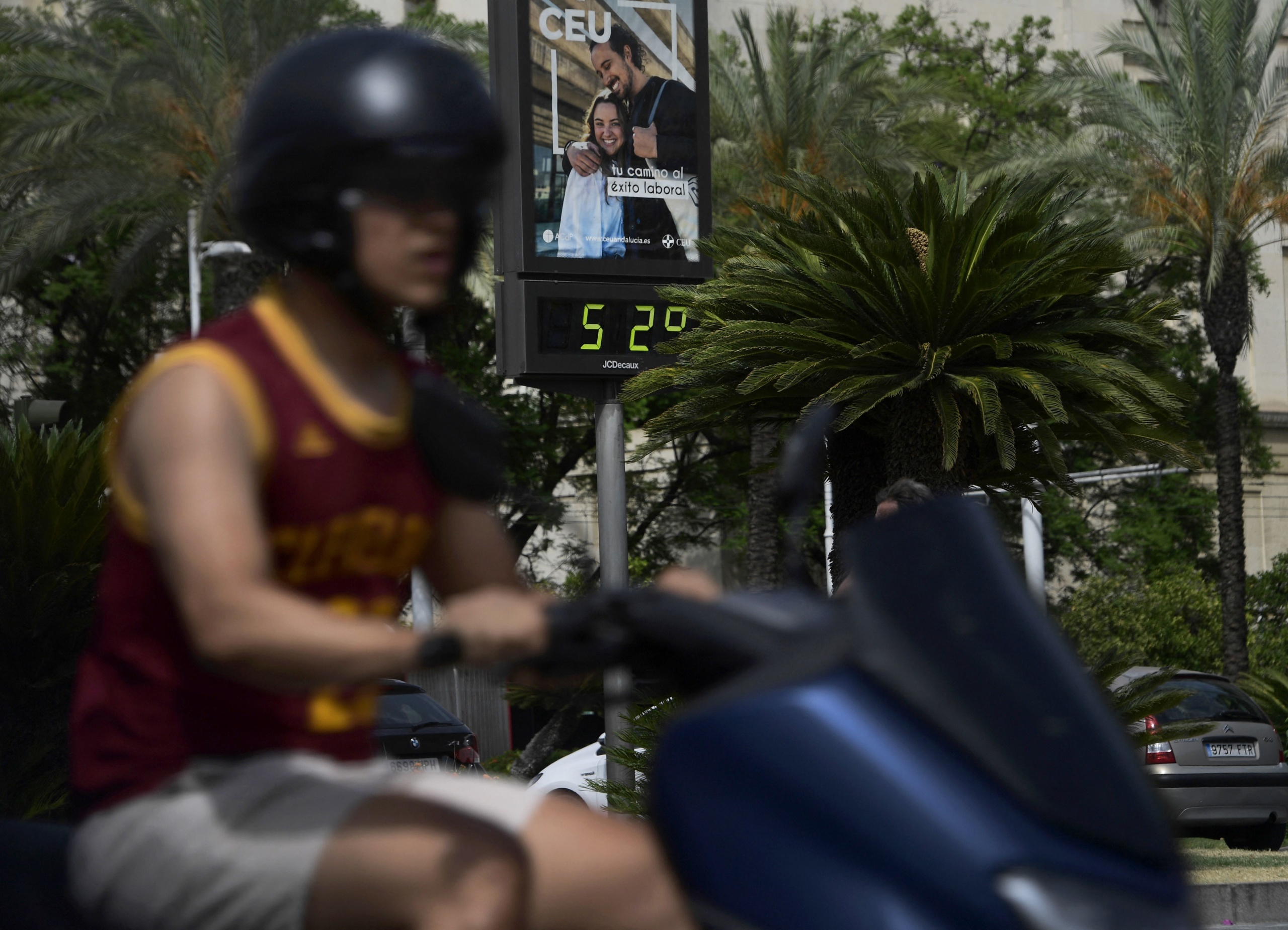 52 fokot mutat az utcai hőmérő Sevillában, előtérben egy robogós.