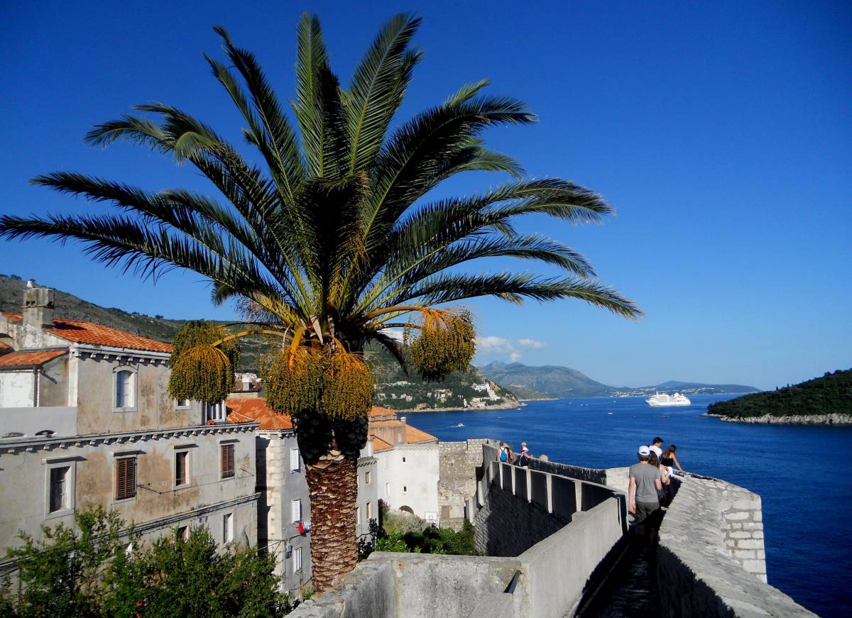 Turisták sétálnak az óvárost körülölelő, újjáépített várfalon. Jobbra a távolban egy utasszállító hajó halad el az Adriai-tengeren Lokrum szigete mellett.