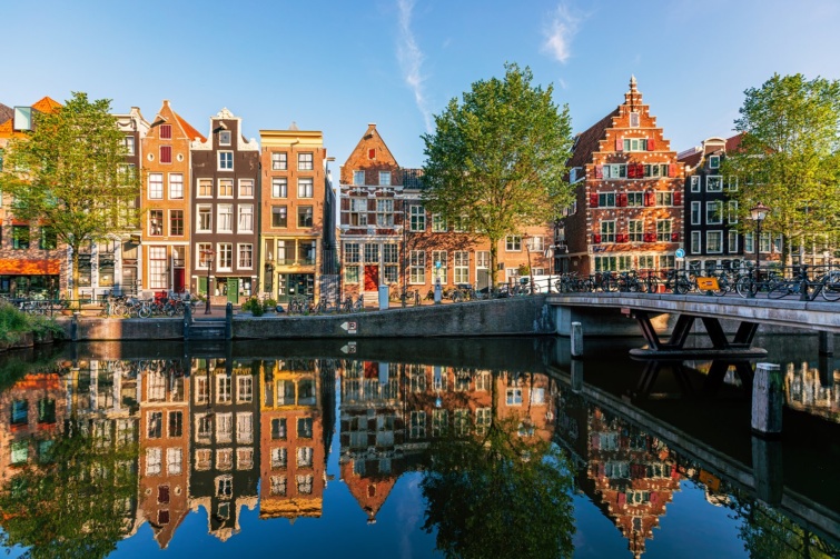 Egy holland város látható a képen, vízparttal. A vízben tükröződnek a szép, színes holland stílusban épült házak. 