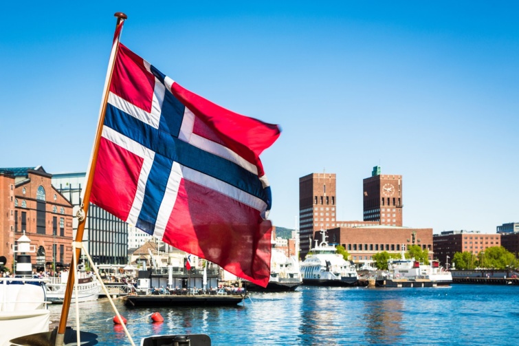 A norvég zászló lobog a szélben, a háttérben vízpart és néhány épület.