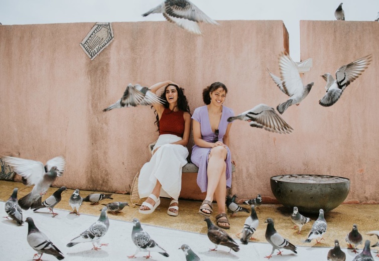 Két boldog olasz nő látható a képen, a nők röpködő galambok között ülnek egy krémszínű fal előtt nevetve.