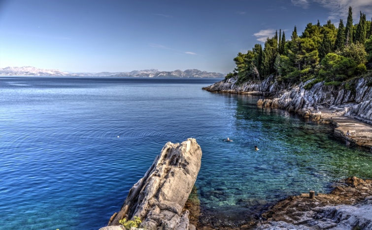 Gyönyörű horvát, sziklás tengerpart, átlátszó kék víz és kék ég, a kép oldalán örökzöld növények övezik a partot.