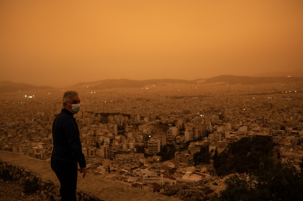 Maszkos férfi áll egy hegy tetején Athén fölött és nézi a szaharai portól narancsba borult várost.