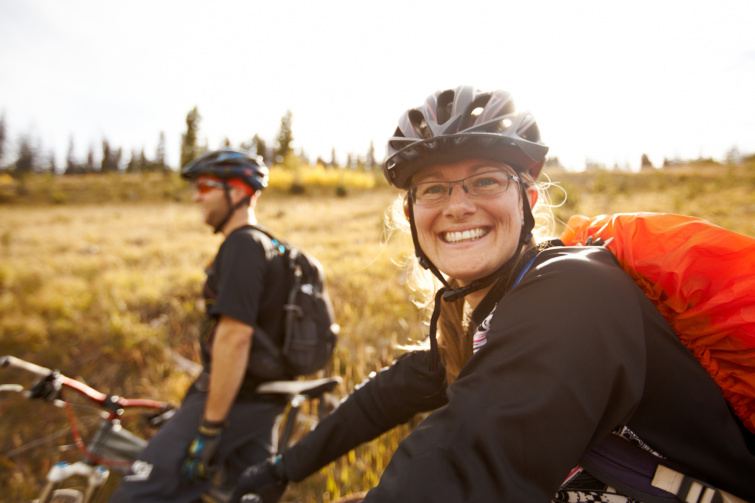 Egy bicikliző pár, az előtérben szőke nő mosolyog, bukósisakkal, piros táskával, körülöttük a természet