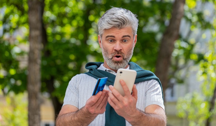 Egy meglepődött kozépkorú férfi egy bankkártyát és egy mobiltelefont tart a kezében az utcán.