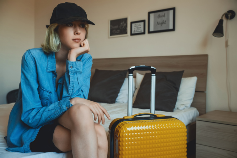 borond - Startlap Utazás Fiatal lány baseball sapkában sárga bőrönddel ül az ágy szélén