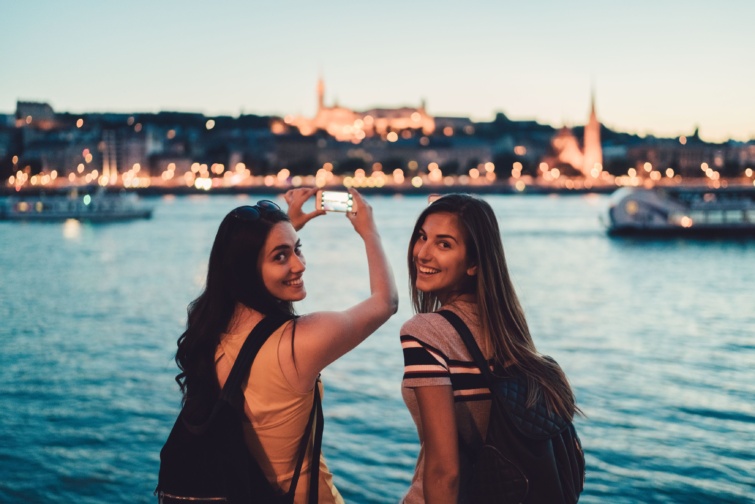 turistak budapesten - Startlap Utazás Két lány áll a pesti Dunaparton, egyikőjük fotózik.