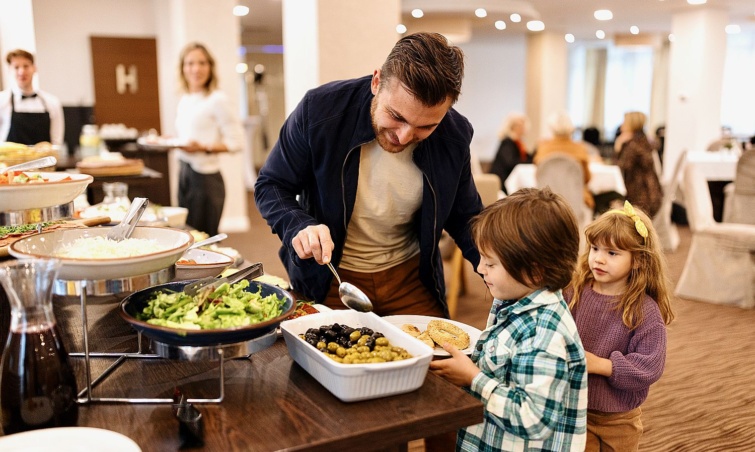 Family enjoying breakfast buffet together in the hotel - Startlap Utazás Egy férfi ételt rak a szállodai svédasztalról két kisgyerek tnyérjába.