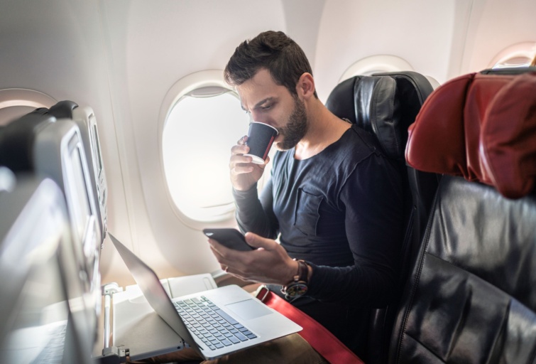 Férfi kávét iszik, a telefonját és a laptopját is használja a repülőgépen.