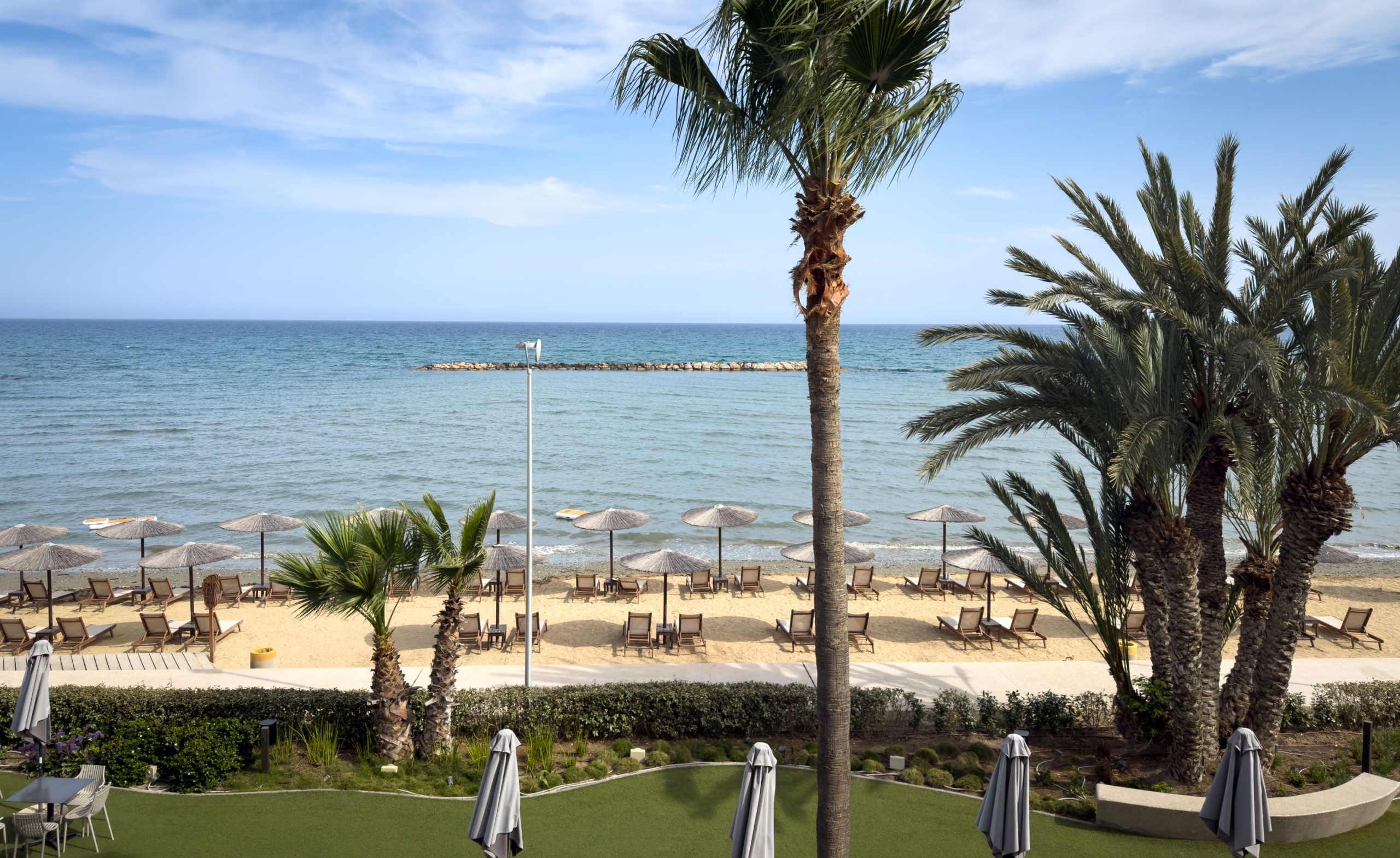 A Mercure Larnaca Beach Resort erkélyéről a szálloda pálmafákkal, napozóágyakkal, napernyőkkel és a Földközi-tengerrel teli strandja látható Orokliniben, a Larnaca kerületben