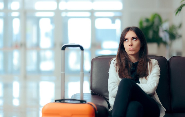 Bored Woman with Suitcase in Airport Waiting Room - Startlap Utazás Csalódott fiatal nő ül egy váróteremben, mellete narancssárga bőrönd.
