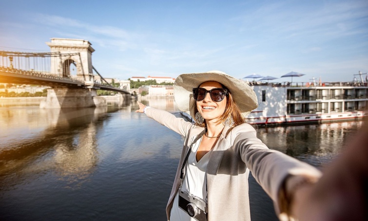 Napszemüveges, kalapos fiatal nő mosolyogva a Dunára mutat, háttérben egy hajó és a Lánchíd.