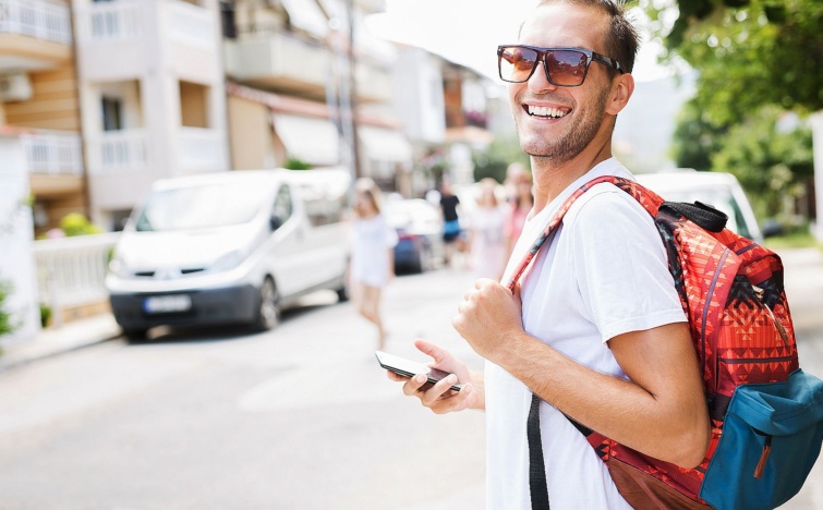Young man on the street with a smartphone - Startlap Utazás Fiatal, napszemüveges, hátizsákos, kezében mobiltelefont tartó férfi nevet az utcán.