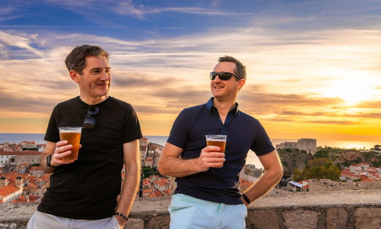 Two male tourist friends enjoying a beer in old town Dubrovnik Croatia - Startlap Utazás Két középkorú férfi kezében sör, háttérben egy tengerparti város és a naplemente