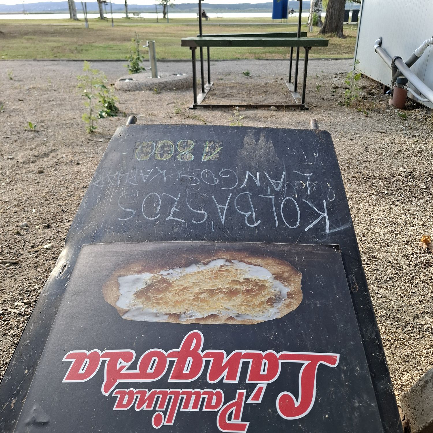 Egy büfé lángost reklámozó, ledöntött hirdetőtáblája egy strandon.
