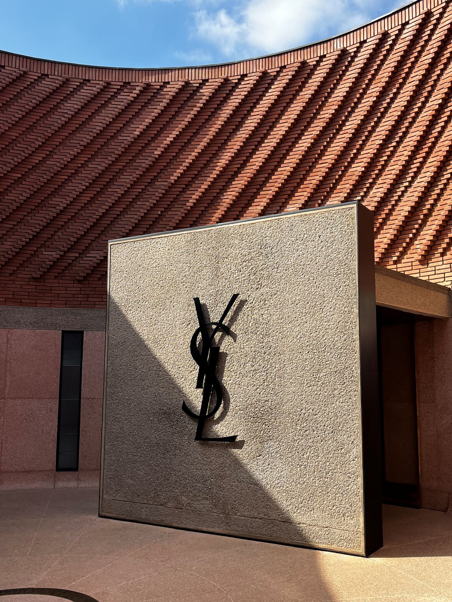 A képen az Yves Saint Laurent múzeum előcsarnoka látható. Vörös tégla falak veszik körül a logót ábrázoló betonfalat.