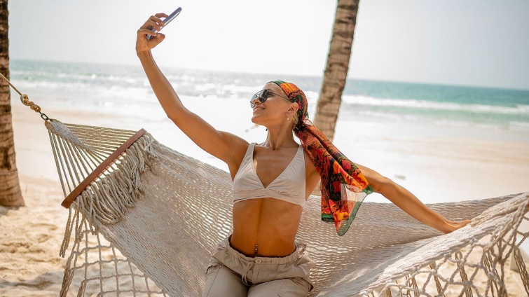 Stylish young woman relaxing in a hammock on a tropical beach taking a joyful selfie with the ocean behind her Dressed in beachwear she enjoys a carefree moment under the sun - Startlap Utazás Egy tengerparti függőágyban ülő fiatal nő szelfizik.