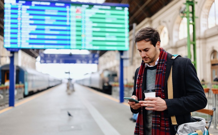 Young businessman using his phone at a railroad station - Startlap Utazás Fiatal férfi mobiltelefonozik egy vasútállomáson.