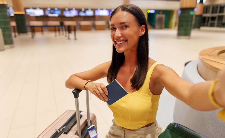 Happy woman at airport taking selfie while sitting with luggage and holding passport capturing moment waiting for her flight to depart - Startlap Utazás Fiatal, mosolygó nő útlevéllel a kezében és bőrönddel egy repülőtéren.