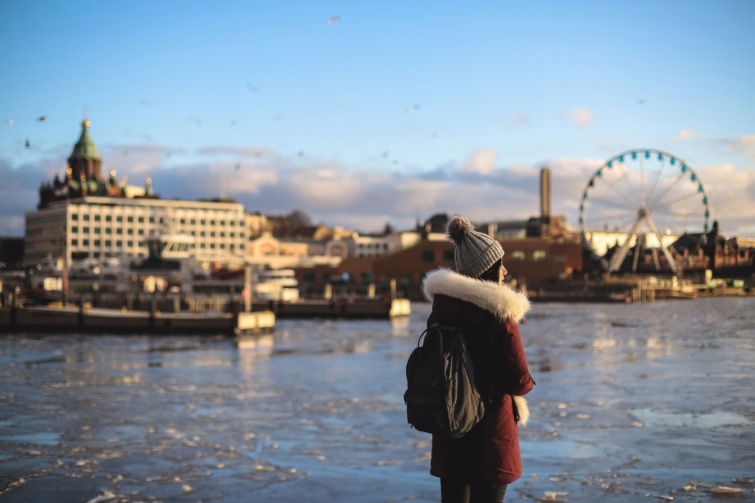 helsinki - Startlap Utazás Fiatal nő áll a SkyWheel Helsinki előtt; a háttérben – hajnalban – az Uspenski ortodox székesegyház és Alvar Aalto Enso-Gutzeit-székháza látszik.