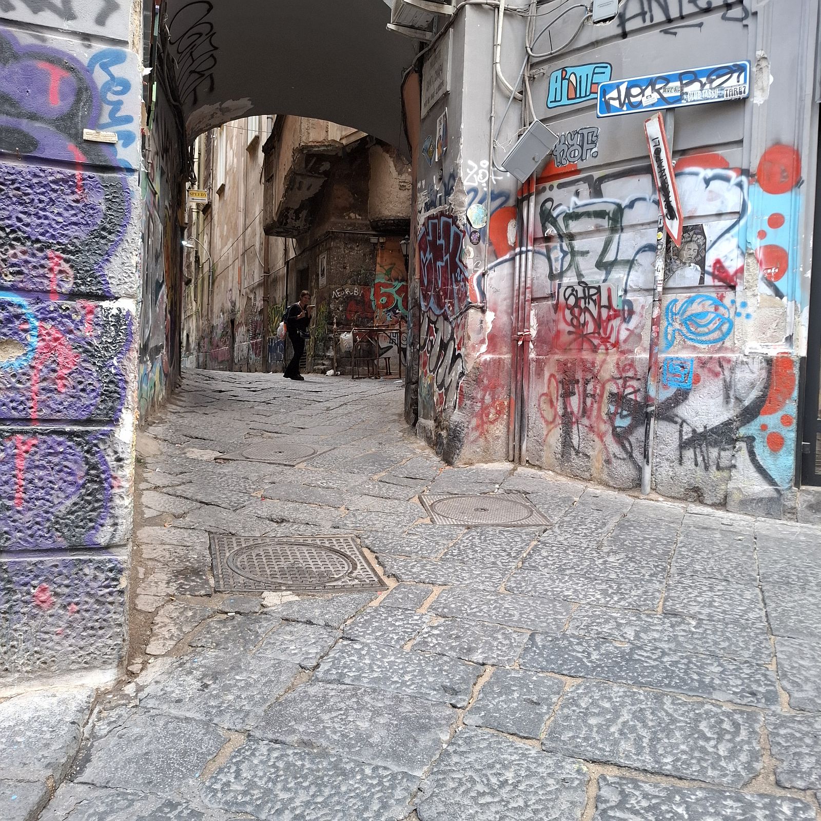 Nápoly történelmi belvárosának graffitikkel borított, szűk sikátora, a kőburkolatú utcán egy gyalogossal.