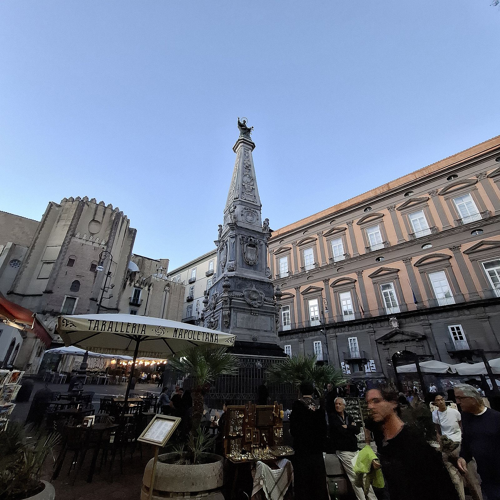 Nápoly történelmi belvárosában, a Piazza San Domenico Maggiore téren álló díszes barokk obeliszk, háttérben narancsszínű homlokzatú palotával és középkori épületekkel. Az előtérben szabadtéri vendéglátóhelyek és turisták láthatók a kék ég alatt.