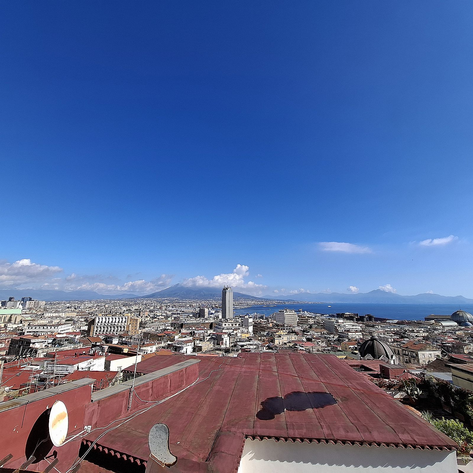 Panorámakép Nápoly városáról, az előtérben vörös tetőkkel, a háttérben a város sűrűn beépített házaival, a Nápolyi-öböl kék vizével és a távolban magasodó hegyekkel, derült, felhős égbolt alatt.