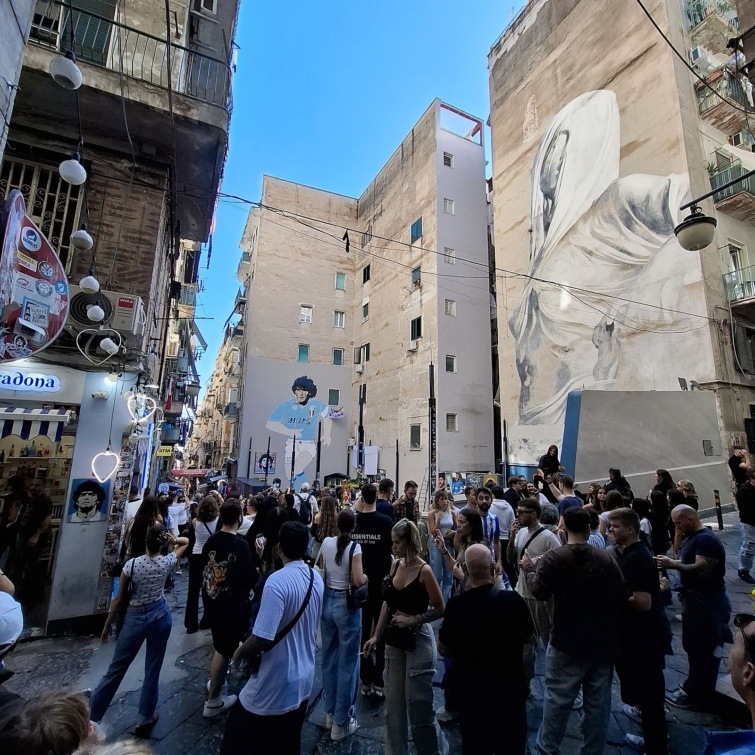 napoly-12 - Startlap Utazás Nápoly szűk utcájában turisták tömege gyűlik össze a Diego Maradonát ábrázoló falfestmények előtt. A magas, kopott homlokzatokon hatalmas portré és egy világoskék Napoli-mezben futballozó Maradona alakja látható, az utcát pedig ajándékboltok, erkélyek és vezetékek szegélyezik.