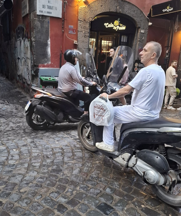 napoly-18 - Startlap Utazás Nápolyi hangulatú, macskaköves utcán robogókon ülő férfiak egy olasz étterem bejárata előtt. A háttérben vörös falak, graffitik és meleg fényű éttermi felirat látható.