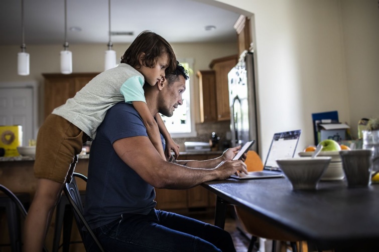 Egy férfi home office-ban a telefonját nézi, egy gyerek átöleli a nyakát.