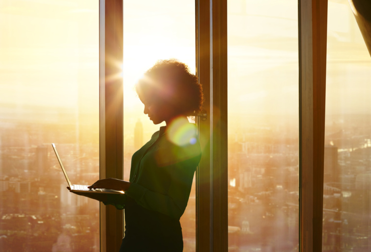 Egy lány napsütötte ablaknál állva laptopozik