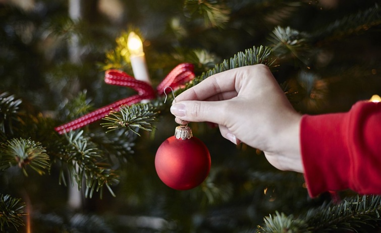 Egy kéz egy piros díszgömböt aggat a karácsonyfa ágára.