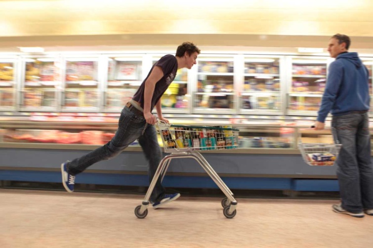 Egy élelmiszerboltban egy férfi bevásárlókocsira támaszkodva gurul, egy másik nézi.