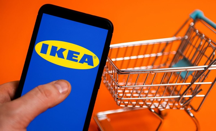 IKEA Startlap Vásárlás IKEA felirat egy okostelefon kijelzőjén, a háttérben egy bevásárlókocsi.
