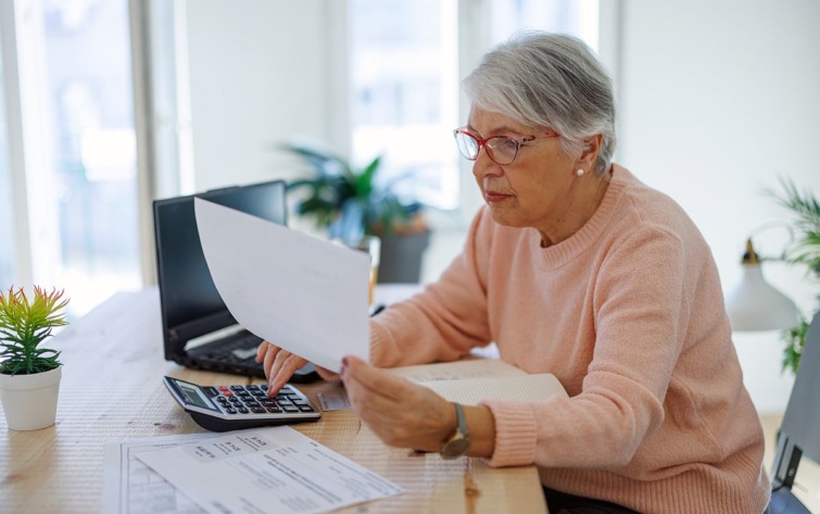 Egy nyugdíjas nő egy iratot olvas és számológépen számol.