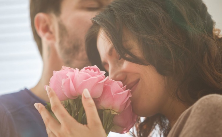 Egy párjához simuló, mosolygó nő beleszagol a kezében tartott rózsacsokorba.
