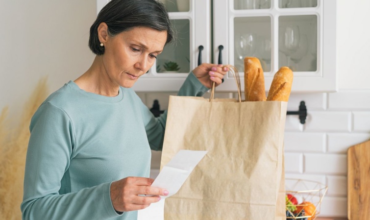 Egy nő otthon vásárlás után a blokkot nézi, másik kezében egy papírzacskó, benne kenyér.