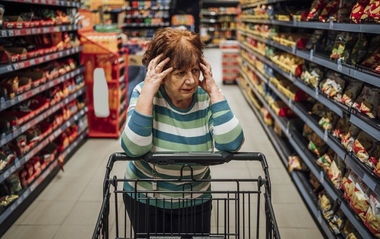 Egy idősebb, gondterhelt nő a bevásárlókocsira támaszkodba a bolt kínálatát nézi.