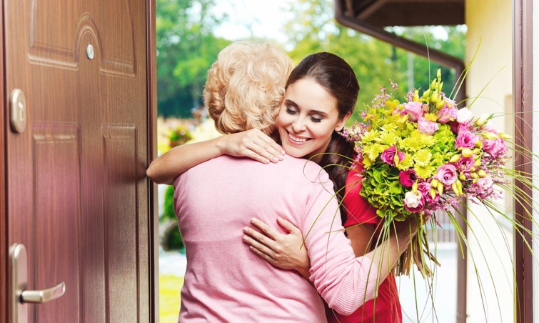 Egy fiatal nő és egy virágcsokrot a kezében fogó idősebb nő megöleli egymást az ajtóban.