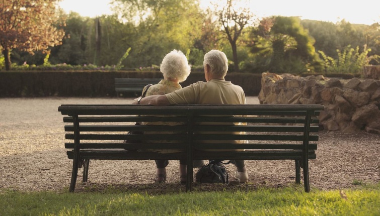 Egy nyugdíjas pár egy padon ül a fotósnak háttal egy parkban.