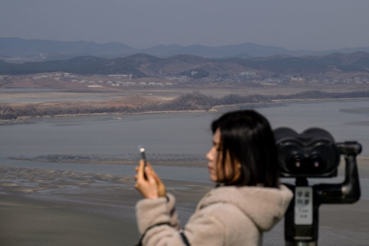 Egy látogató a telefonját használja a koreai demilitarizált övezet észak-koreai oldalának látképe előtt 