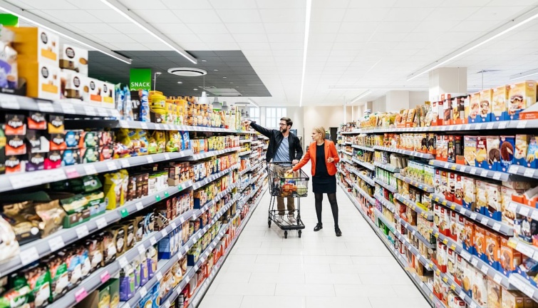 Egy bevásárlókocsit toló középkorú pár válogat a termékek között egy élelmiszerboltban.