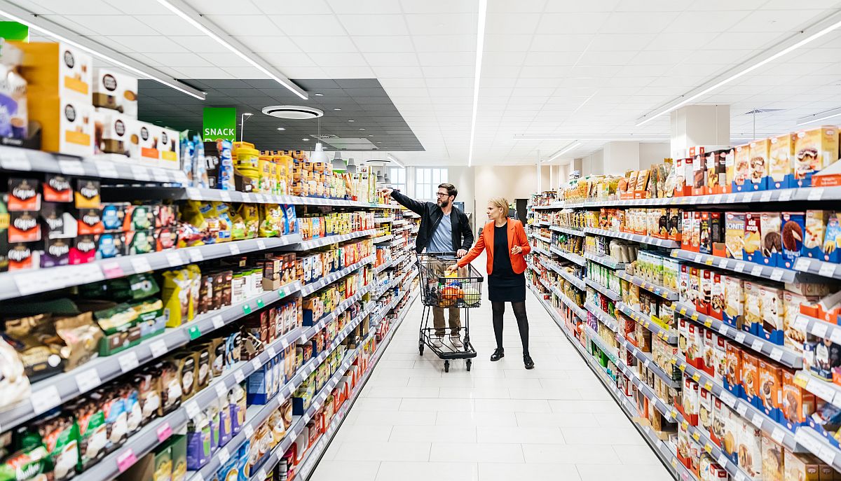 Egy bevásárlókocsit toló középkorú pár válogat a termékek között egy élelmiszerboltban.